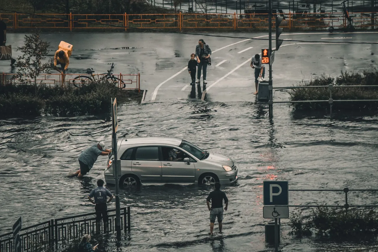 A small silver car stalled in brown floodwater at a city crossing while people wade across the street