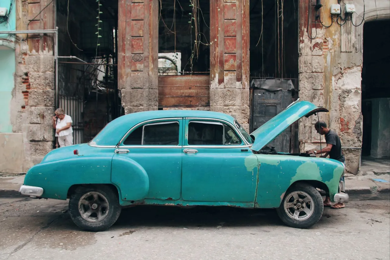 An old teal sedan parked on a city street with its hood raised while a man leans in over the engine