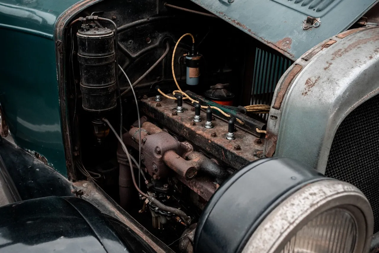 Open hood of an old car showing a rusted engine block, hoses and a round headlight
