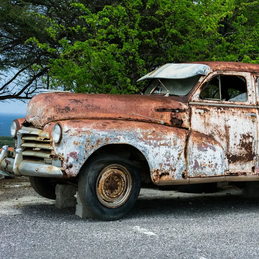 A rusted 1940s-style sedan with faded paint, raised on blocks in an asphalt lot in front of trees